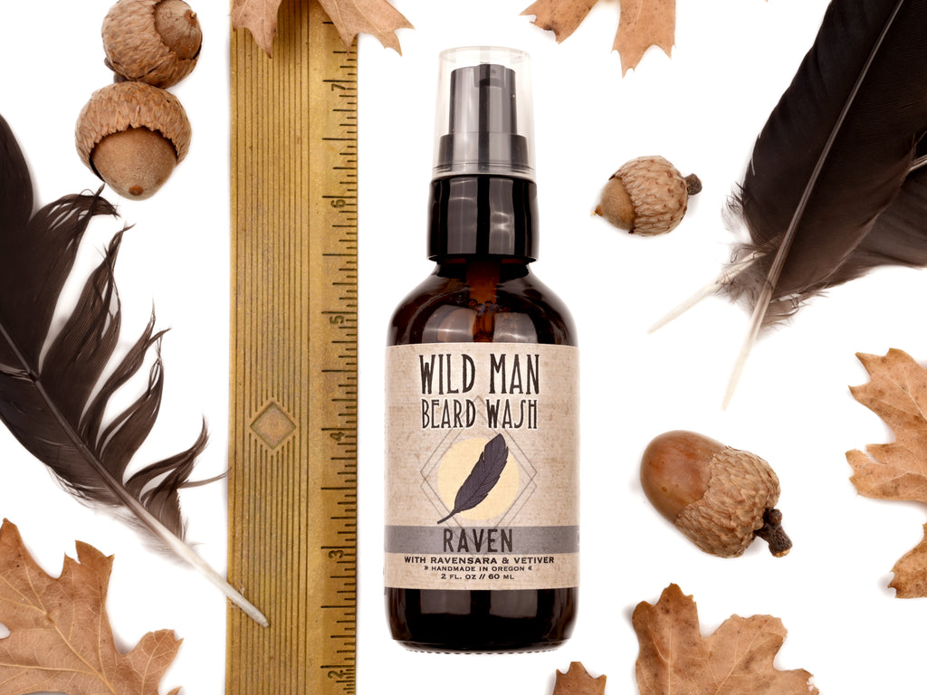 Wild Man Beard Wash - Raven scent in 2oz amber glass bottle. Shown next to ruler at about 5" tall. Black feathers, acorns and dried oak leaves surround. 