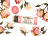 Wild Rose Rosebud Botanical Lip Balm in a biodegradable tube on a white background with dried roses surrounding. The cap is removed revealing a creamy pink balm.