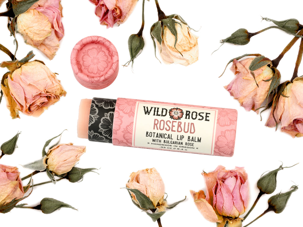Wild Rose Rosebud Botanical Lip Balm in a biodegradable tube on a white background with dried roses surrounding. The cap is removed revealing a creamy pink balm.