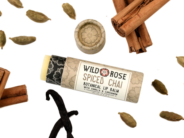 Wild Rose Spiced Chai Lip Balm in a biodegradable tube on a white background with cinnamon sticks, cardamom pods and vanilla bean. The cap is off revealing a creamy white balm.