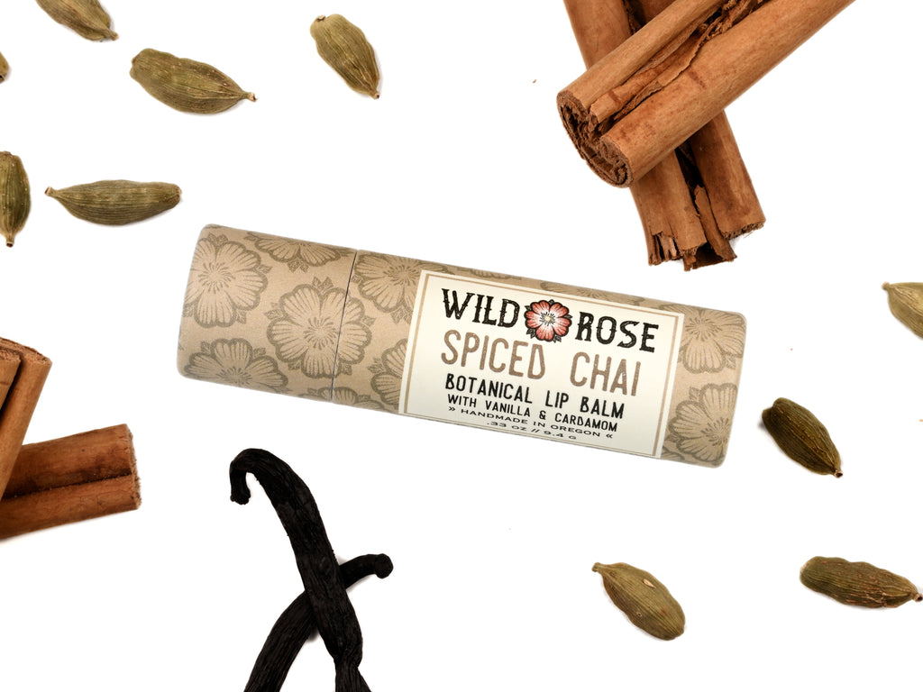 Wild Rose Spiced Chai Lip Balm in a biodegradable tube on a white background with cinnamon sticks, cardamom pods and vanilla bean. 