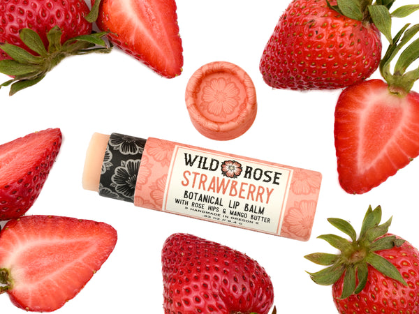 Wild Rose Strawberry Botanical Lip Balm in a biodegradable tube on a white background with fresh strawberries surrounding. The cap is removed revealing a creamy pink balm.