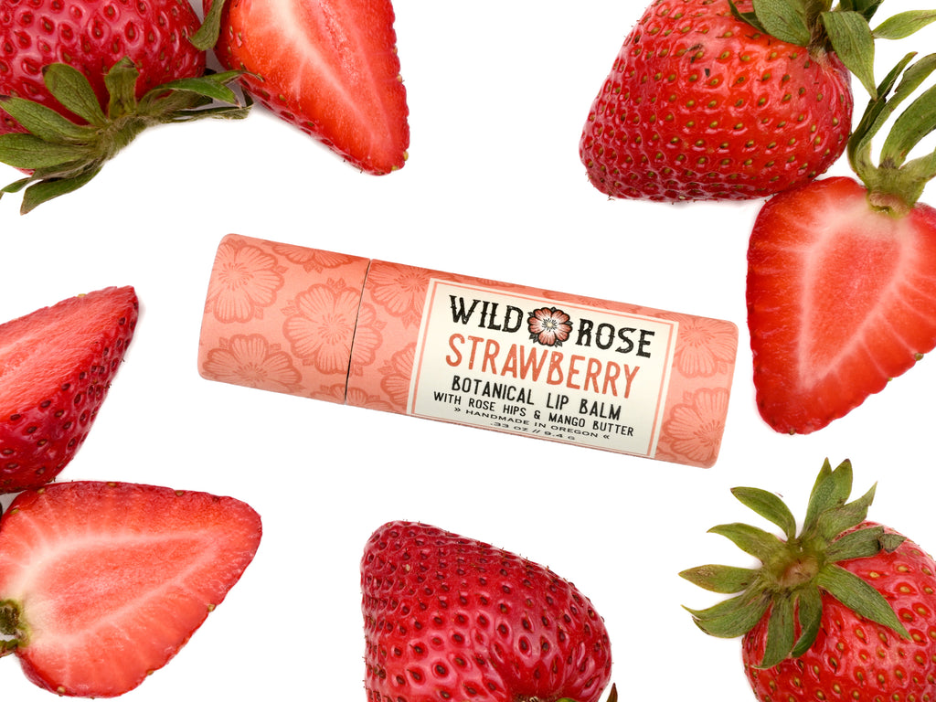 Wild Rose Strawberry Botanical Lip Balm in a biodegradable tube on a white background with fresh strawberries surrounding. 
