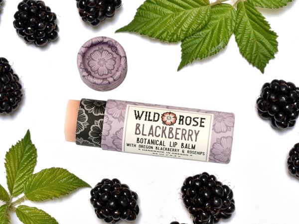 Blackberry Natural Lip Balm in a biodegradable paper tube. The cap is off revealing a creamy soft pink balm. Blackberries and leaves surround.