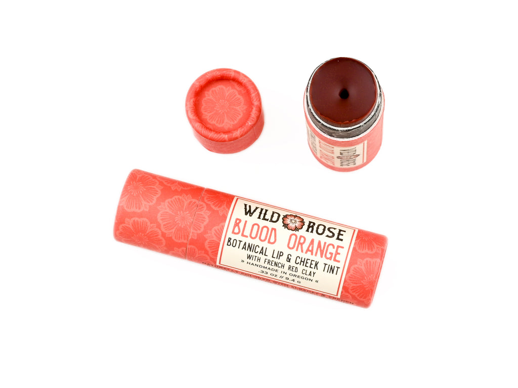 Wild Rose Blood Orange Botanical Lip & Cheek Tint shown with its coral pink patterned tube packaging, a removed cap, and an open jar displaying the dark reddish-brown tinted product inside, on a solid white background.