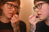 A person with glasses and tattoos applies Wild Rose Blood Orange Lip Tint while looking into a mirror, showing both a front and side profile view.