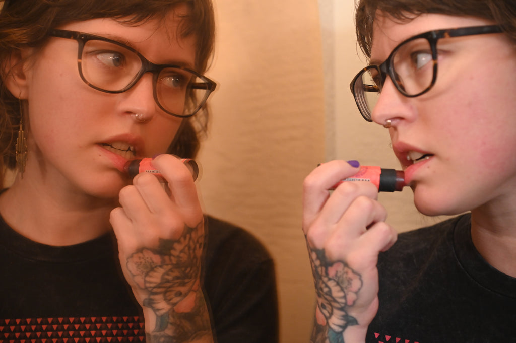 A person with glasses and tattoos applies Wild Rose Blood Orange Lip Tint while looking into a mirror, showing both a front and side profile view.