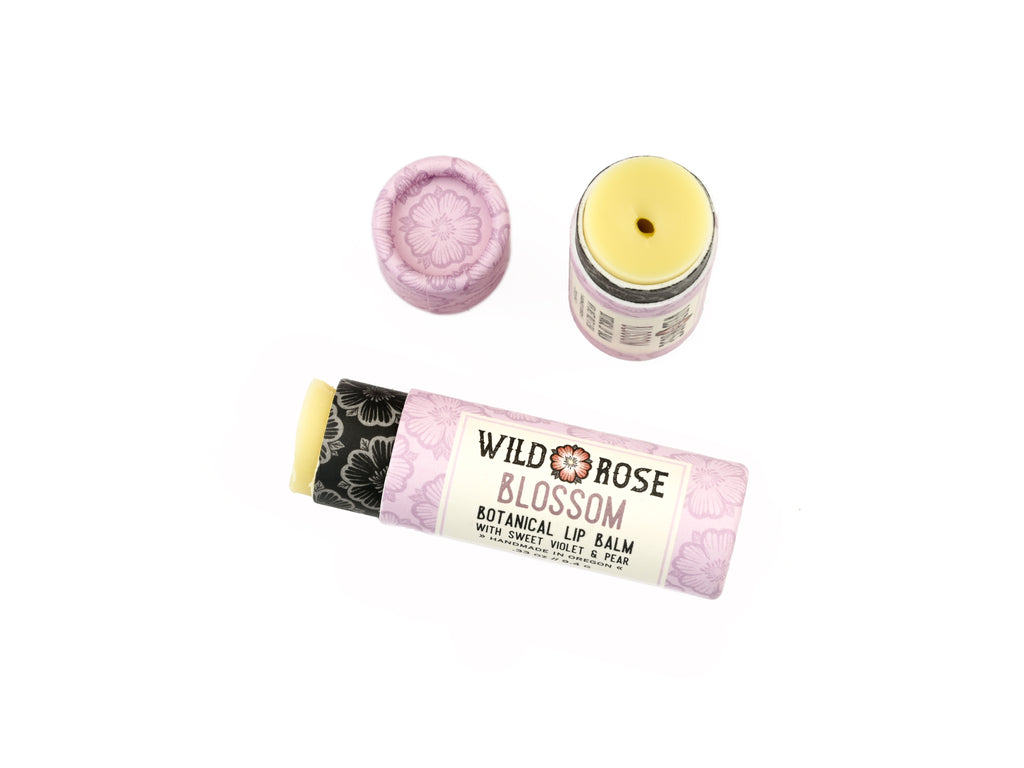 Wild Rose Blossom botanical lip balm shown with its light purple floral tube packaging on a white background. The lid is removed revealing a creamy, light green balm.