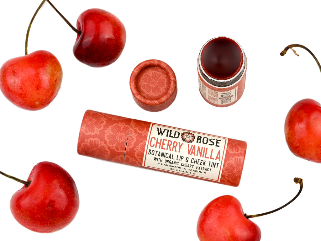 Wild Rose Cherry Vanilla Botanical Lip & Cheek Tint in a biodegradable tube, surrounded by fresh cherries on a white background.