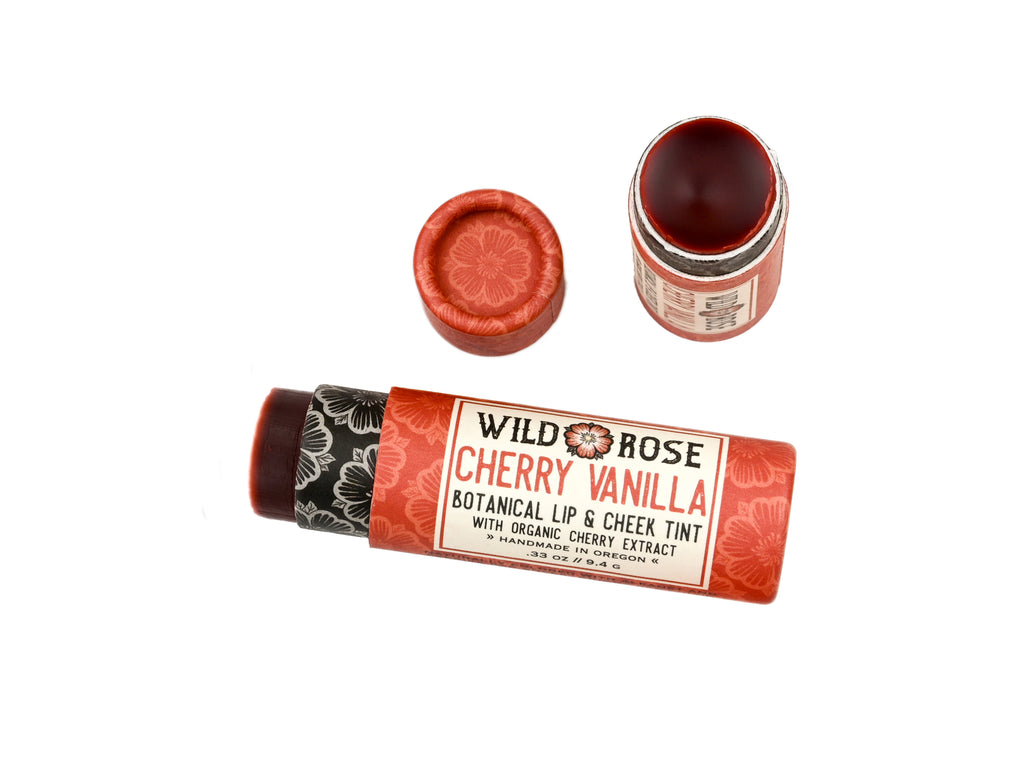 Wild Rose Cherry Vanilla Botanical Lip & Cheek Tint in a biodegradable tube on a white background. The cap is removed revealing a dark red creamy tinted balm.