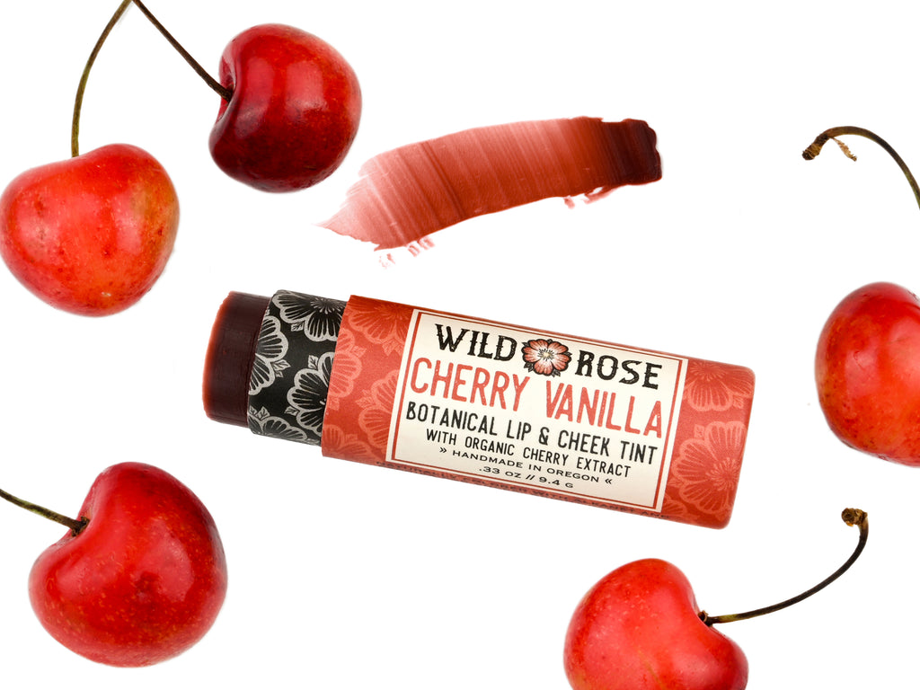 Wild Rose Cherry Vanilla Botanical Lip & Cheek Tint in a biodegradable tube on a white background with fresh cherries surrounding. The cap is removed revealing a dark red creamy tinted balm.