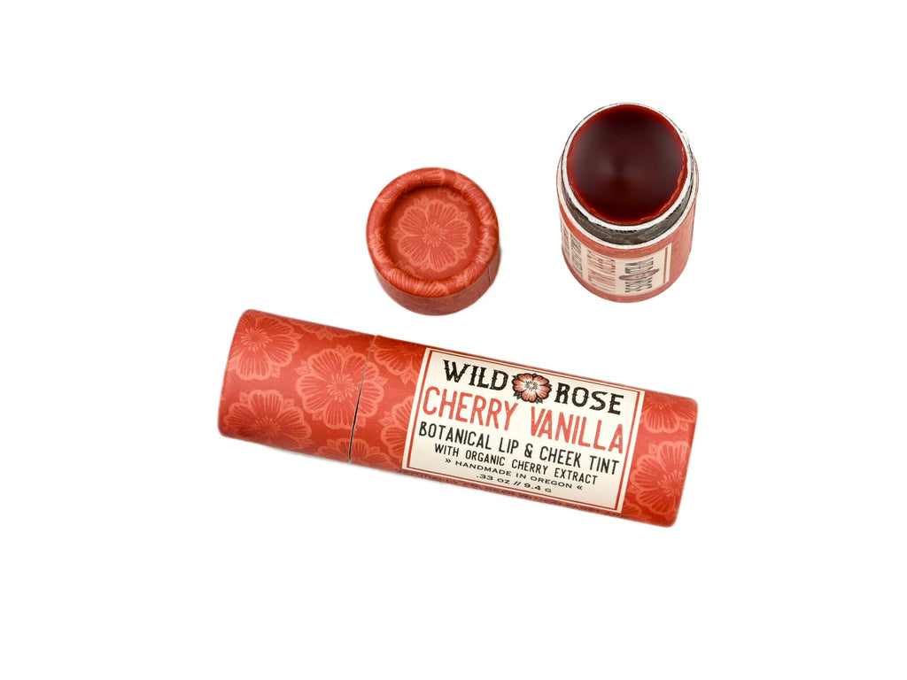 Wild Rose Cherry Vanilla Botanical Lip & Cheek Tint in a biodegradable tube on a white background. The cap is removed revealing a dark red creamy tinted balm.