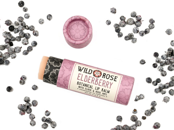 Elderberry Natural Lip Balm in a biodegradable paper tube with elderberries scattered around. The cap is off revealing a creamy, light pink balm.