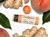 Wild Rose Ginger Peach Botanical Lip Balm in a biodegradable tube, surrounded by fresh peaches, ginger root and green leaves on a white background. The cap is off revealing a creamy orange balm.
