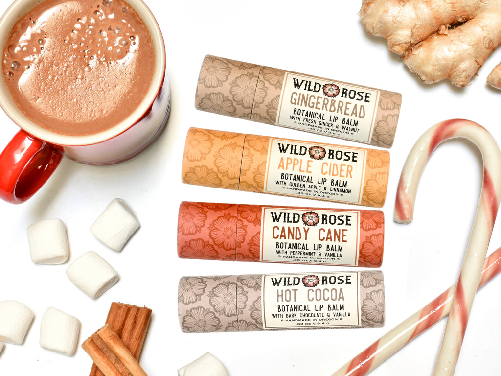 Wild Rose Holiday Lip Balm set with Gingerbread, Apple Cider, Candy Cane and Hot Cocoa Lip Balms in biodegradable tubes. Shown with candy canes, a cup of hot cocoa, marshmallows, ginger root and cinnamon on a white background.