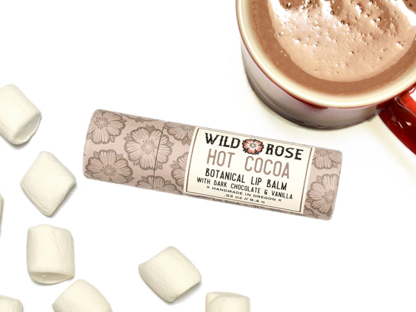 Wild Rose Hot Cocoa Botanical Lip Balm in a biodegradable tube on a white background with marshmallows and a cup of hot cocoa. 