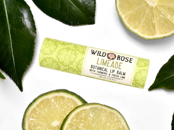 Wild Rose Limeade Botanical Lip Balm in a biodegradable tube on a white background with fresh lime slices and green leaves. 