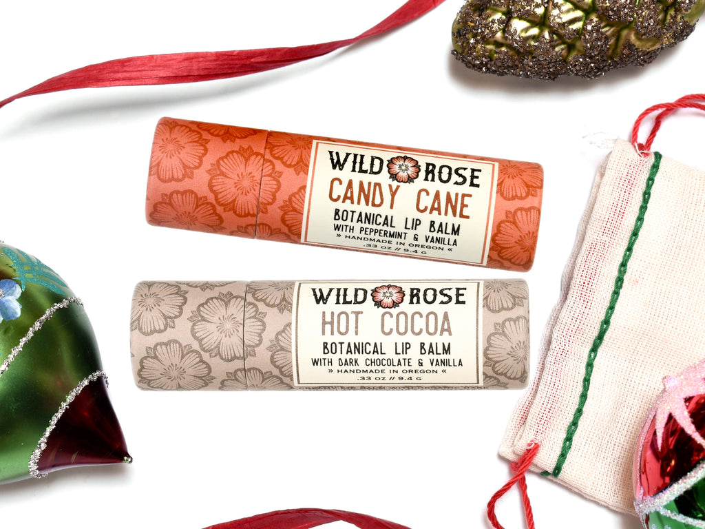 Wild Rose Holiday Lip Balm Stocking Stuffer Set with Candy Cane and Hot Cocoa Lip Balms on a white background. Ornaments and ribbon surround. Shown with a cotton gift bag with red and green stitching.