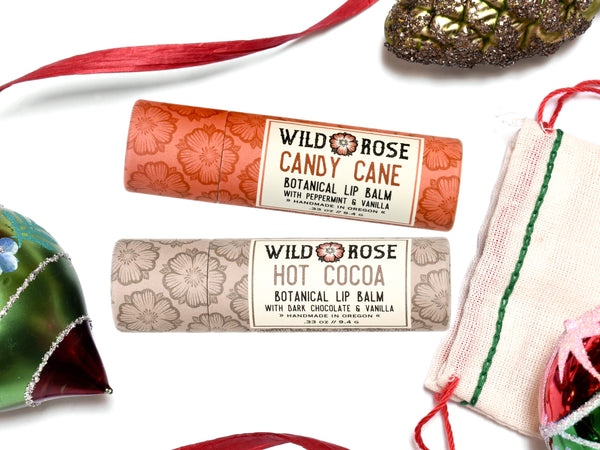 Wild Rose Holiday Lip Balm Stocking Stuffer Set with Candy Cane and Hot Cocoa Lip Balms on a white background. Ornaments and ribbon surround. Shown with a cotton gift bag with red and green stitching.