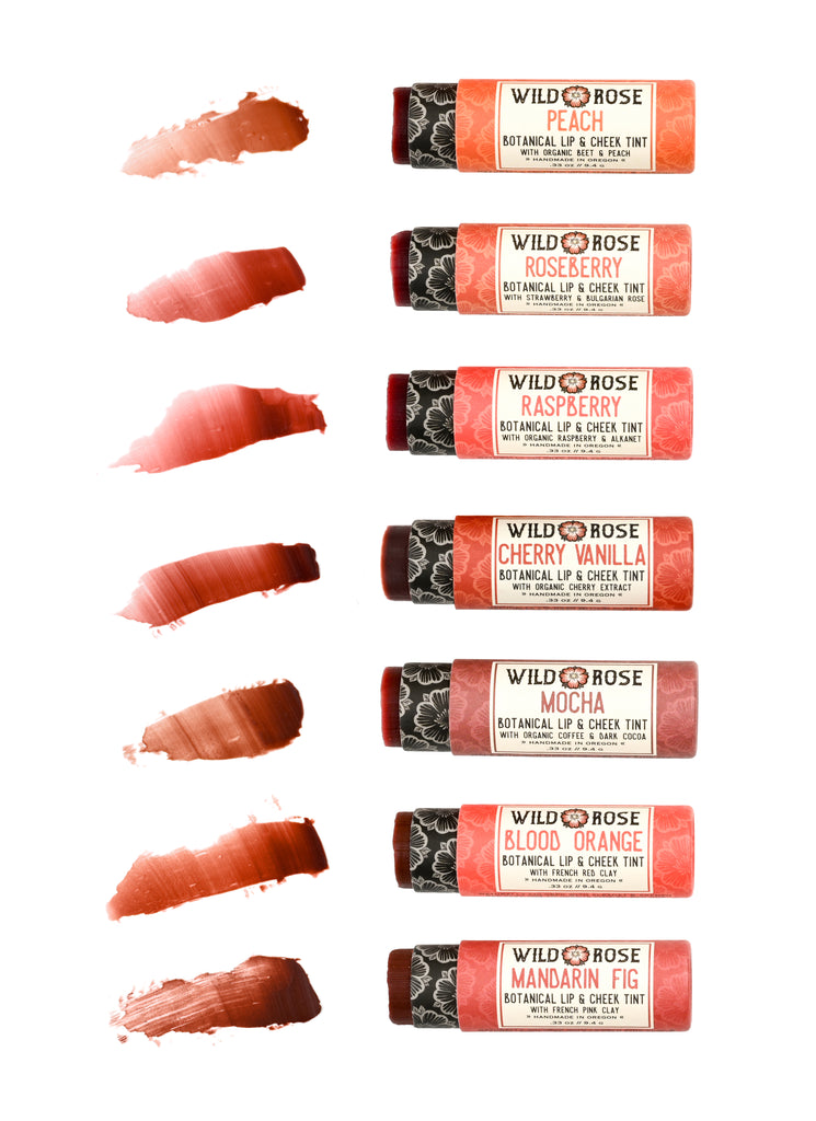 Eight Wild Rose Botanical Lip & Cheek Tint tubes arranged vertically on a white background, each paired with a corresponding textured color smear showing the tints shade, including Peach, Roseberry, Raspberry, Cherry Vanilla, Mocha, Blood Orange, and Mandarin Fig.