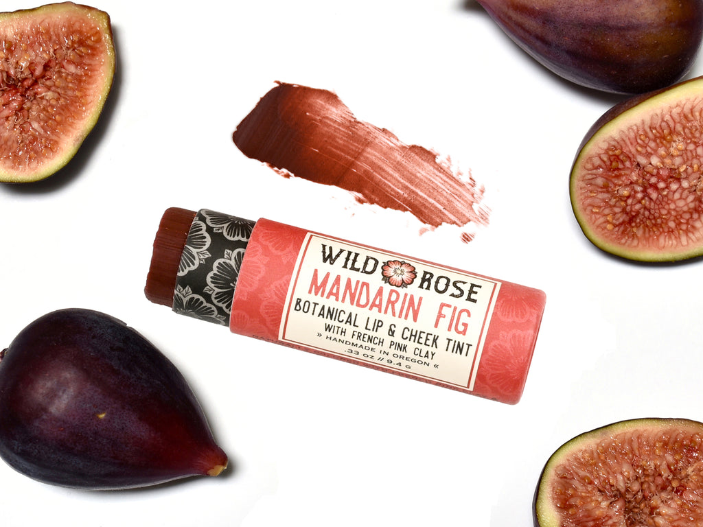 Wild Rose Mandarin Fig Botanical Lip & Cheek Tint in a biodegradable tube on a white background with fresh figs surrounding. The cap is removed revealing a dark red creamy tinted balm.