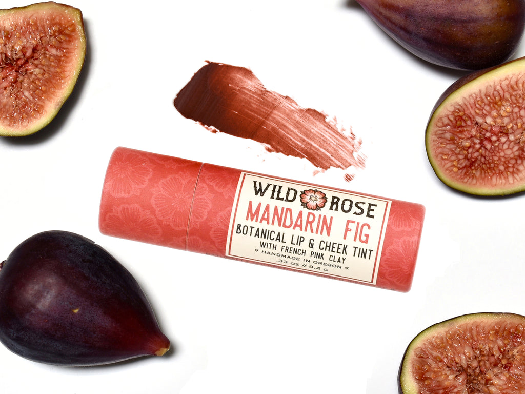Wild Rose Mandarin Fig Botanical Lip & Cheek Tint in a biodegradable tube on a white background with fresh figs surrounding. A color swatch of the tint is above.