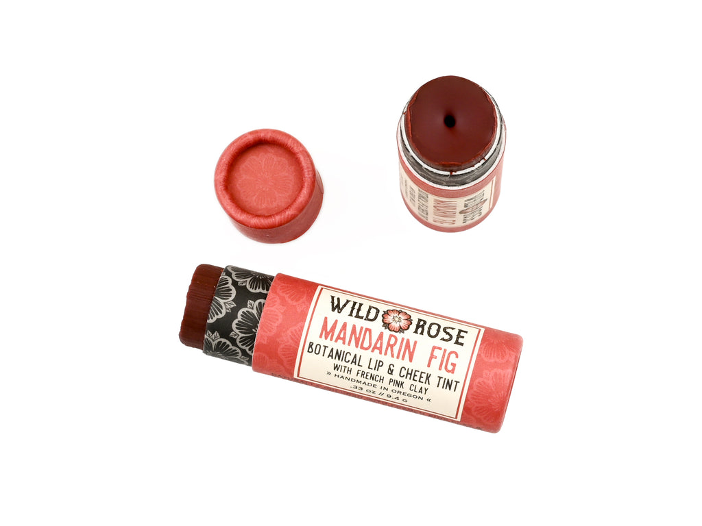 Wild Rose Mandarin Fig Botanical Lip & Cheek Tint in a biodegradable tube on a white background with fresh figs surrounding. The cap is removed revealing a dark red creamy tinted balm.