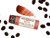 Wild Rose Mocha Botanical Lip & Cheek Tint in a biodegradable paper tube on a white background with coffee beans surrounding. The cap is removed and a color swatch is above, revealing a dark red tint.