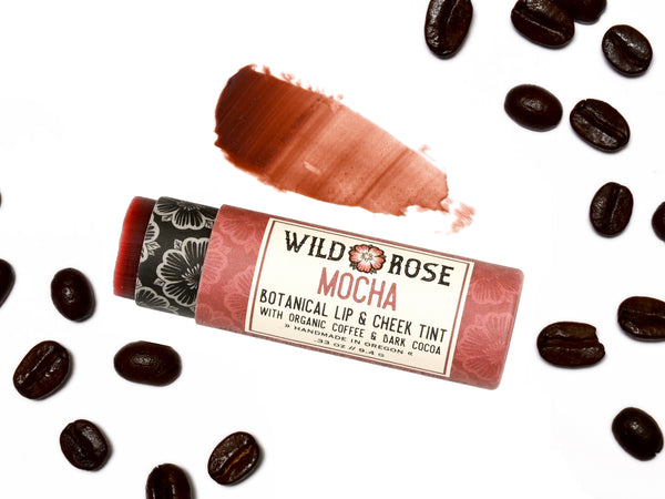 Wild Rose Mocha Botanical Lip & Cheek Tint in a biodegradable paper tube on a white background with coffee beans surrounding. The cap is removed and a color swatch is above, revealing a dark red tint.