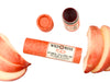 Wild Rose Peach Botanical Lip & Cheek Tint in a biodegradable paper tube on a white background with peach slices surrounding. The cap is removed and a color swatch is above, revealing a red-orange tint.