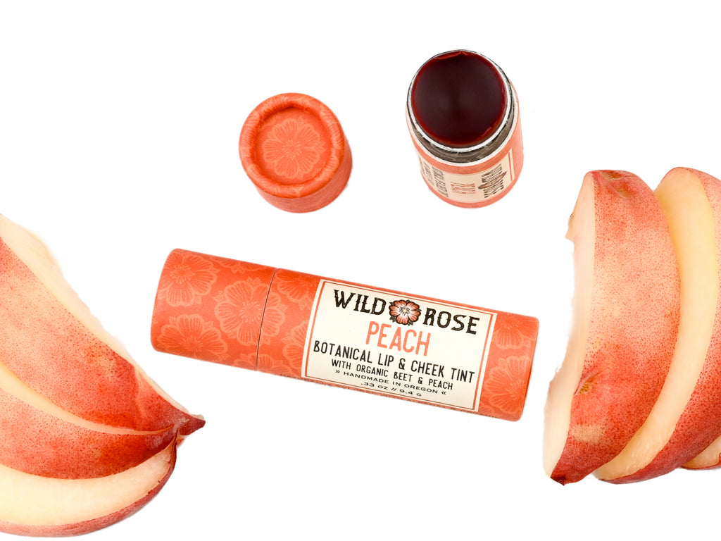 Wild Rose Peach Botanical Lip & Cheek Tint in a biodegradable paper tube on a white background with peach slices surrounding. The cap is removed and a color swatch is above, revealing a red-orange tint.