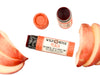 Wild Rose Peach Botanical Lip & Cheek Tint in a biodegradable paper tube on a white background with peach slices surrounding. The cap is removed and a color swatch is above, revealing a red-orange tint.