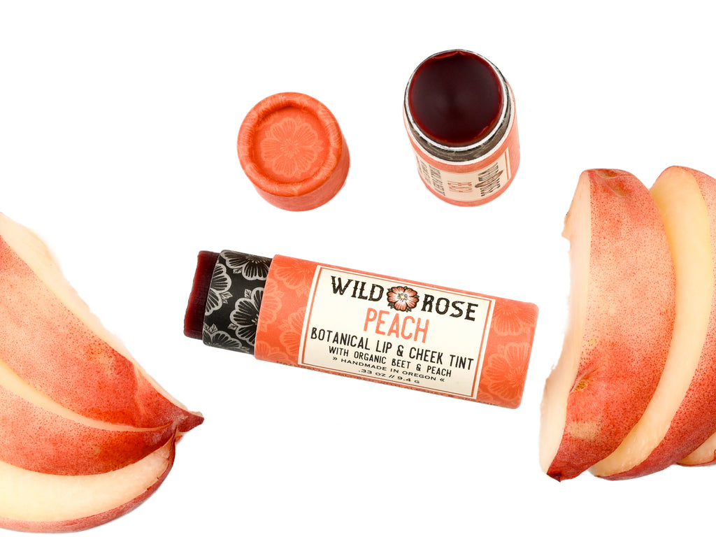 Wild Rose Peach Botanical Lip & Cheek Tint in a biodegradable paper tube on a white background with peach slices surrounding. The cap is removed and a color swatch is above, revealing a red-orange tint.