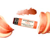 Wild Rose Peach Botanical Lip & Cheek Tint in a biodegradable paper tube on a white background with peach slices surrounding. The cap is removed and a color swatch is above, revealing a red-orange tint.