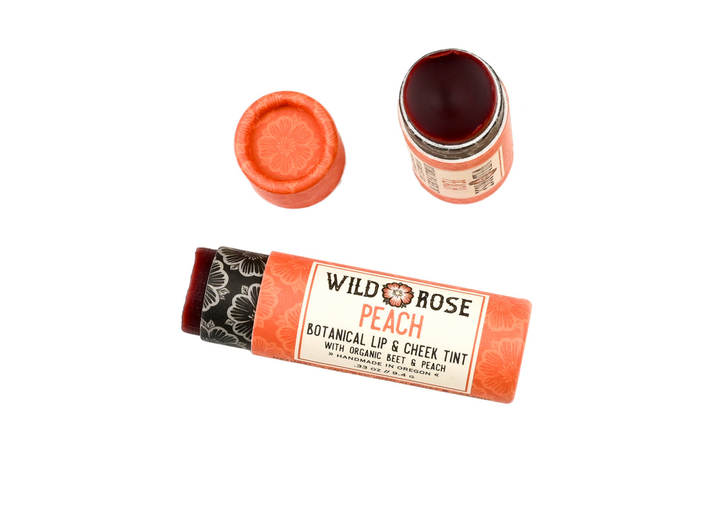 Wild Rose Peach Botanical Lip & Cheek Tint in a biodegradable paper tube on a white background. The cap is removed, revealing a red-orange tint.