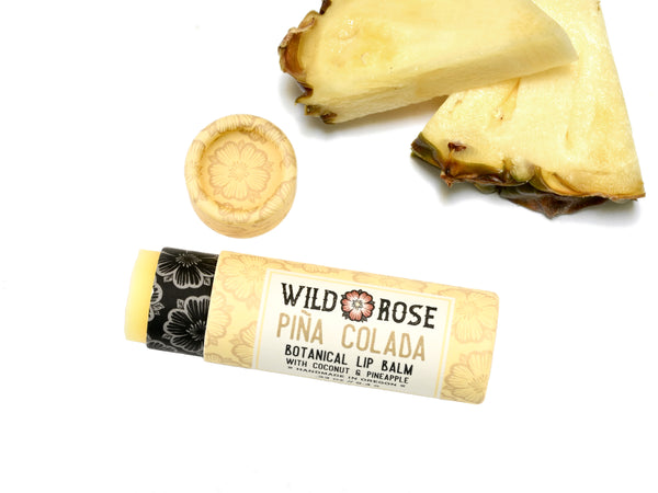 Wild Rose Piña Colada Botanical Lip Balm in a biodegradable tube, surrounded by fresh pineapple on a white background. The cap is off, revealing a creamy yellow balm.