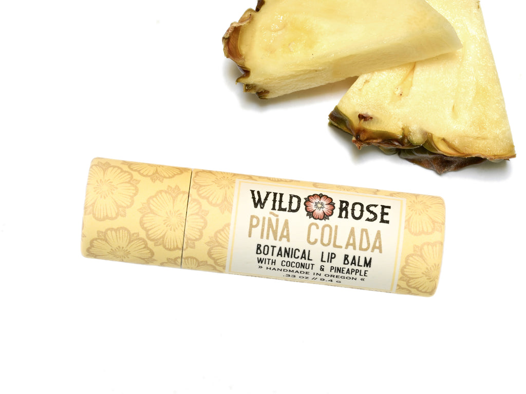 Wild Rose Piña Colada Botanical Lip Balm in a biodegradable tube, surrounded by fresh pineapple on a white background.