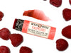 Wild Rose Raspberry Botanical Lip & Cheek Tint in a biodegradable paper tube on a white background with fresh raspberries. The cap is removed and a color swatch is above, revealing a dark red tint.