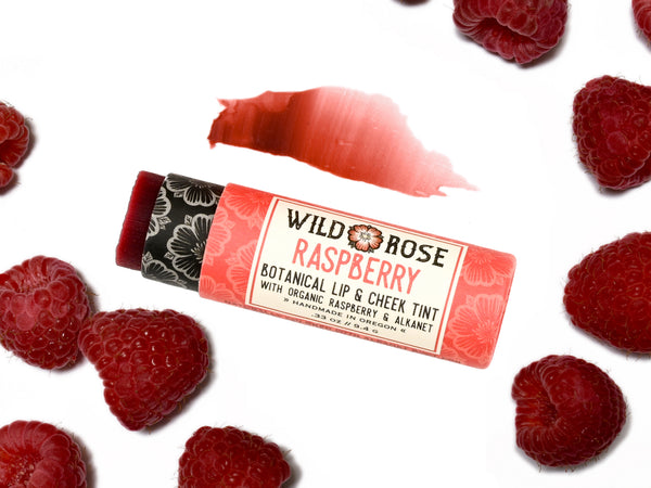 Wild Rose Raspberry Botanical Lip & Cheek Tint in a biodegradable paper tube on a white background with fresh raspberries. The cap is removed and a color swatch is above, revealing a dark red tint.