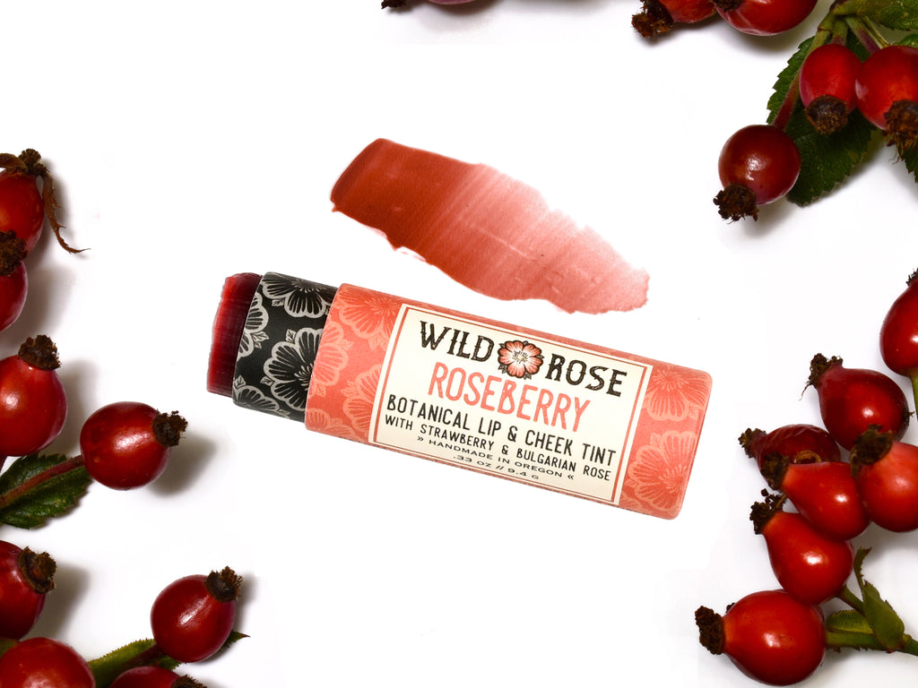 Wild Rose Roseberry Botanical Lip & Cheek Tint in a biodegradable tube, surrounded by rose hips on a white background. The cap is removed revealing a creamy soft red tint with product swatch above.