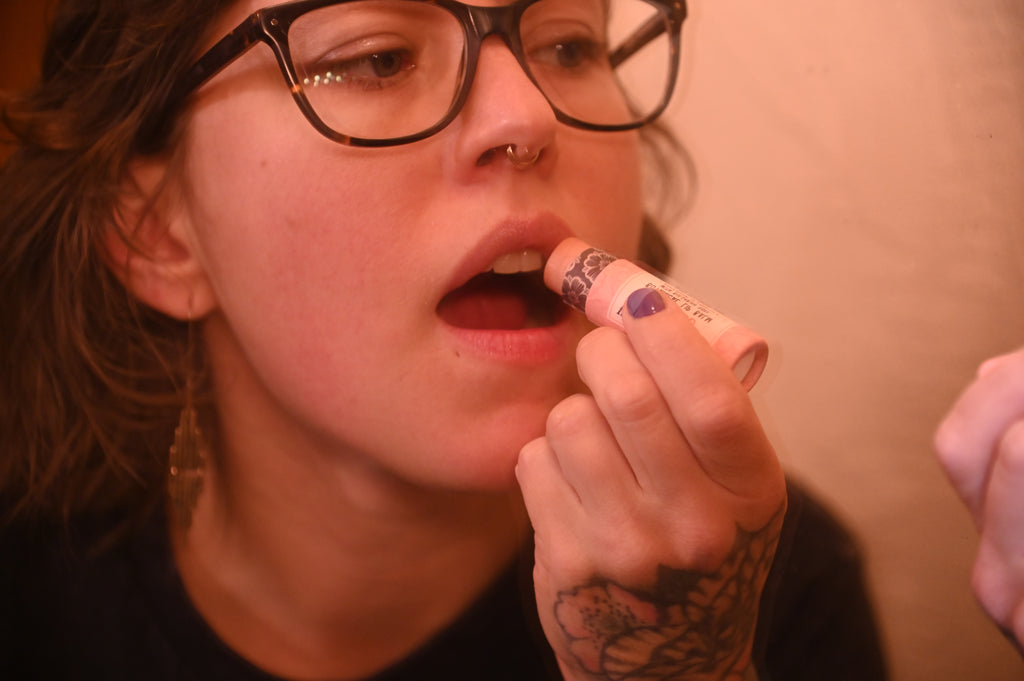 A close-up lifestyle shot of a person with glasses applying Wild Rose Botanical lip balm from a pink biodegradable tube to their lips.