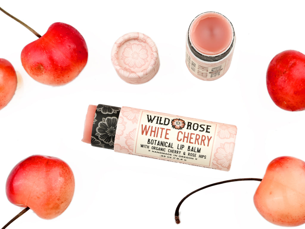 Wild Rose White Cherry Botanical balm in a biodegradable tube shown with the cap removed revealing a creamy pink balm. Surrounded by fresh cherries.