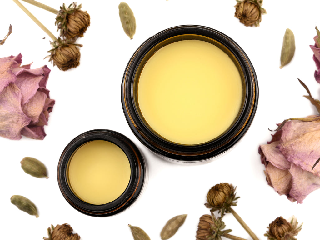 Bloom Gentle Muscle Rub in amber glass jars with lids removed revealing a creamy yellow balm. Dried flowers and cardamom pods surround.