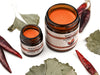 Gingermint Muscle Rub in amber glass jars. The lids are removed revealing a creamy bright orange balm. Dried gingko and peppers surround.