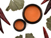 Gingermint Muscle Rub in amber glass jars. The lids are removed revealing a creamy bright orange balm. Dried gingko and peppers surround.