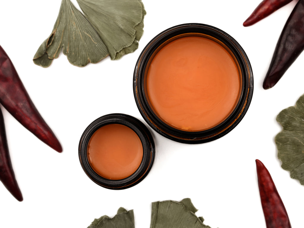 Gingermint Muscle Rub in amber glass jars. The lids are removed revealing a creamy bright orange balm. Dried gingko and peppers surround.