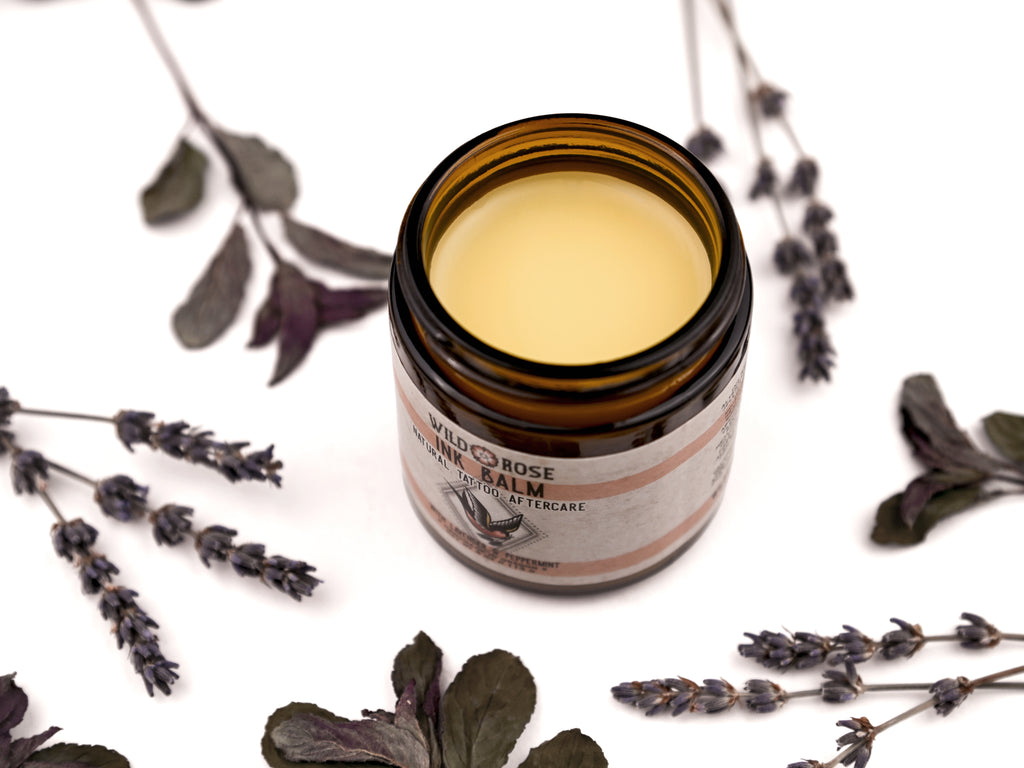 Ink Balm Natural Tattoo Aftercare in an amber glass jar. The lid is removed revealing a creamy yellow balm. Dried lavender and peppermint surround.