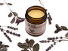 Ink Balm Natural Tattoo Aftercare in an amber glass jar. The lid is removed revealing a creamy yellow balm. Dried lavender and peppermint surround.