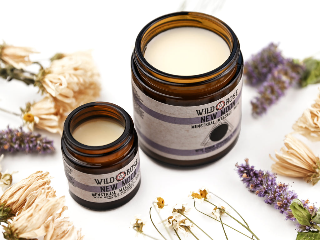 New Moon Menstrual Massage Balm in amber glass jars. The lids are removed revealing a creamy white balm.  Dried flowers surround.